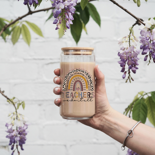 Hand holding a glass mug with a rainbow design and 'Teachers We Do It All' text, surrounded by purple flowers.