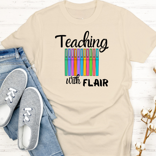 Beige t-shirt with 'Teaching with Flair' text and colorful clip graphic, paired with jeans and sneakers on a light wooden surface.