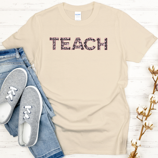 Beige t-shirt with 'TEACH' text, jeans, and sneakers on a white wooden surface.