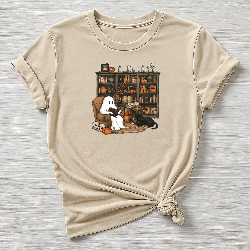 Beige t-shirt with Halloween-themed graphic of a ghost, black cat, and books on a wooden surface.