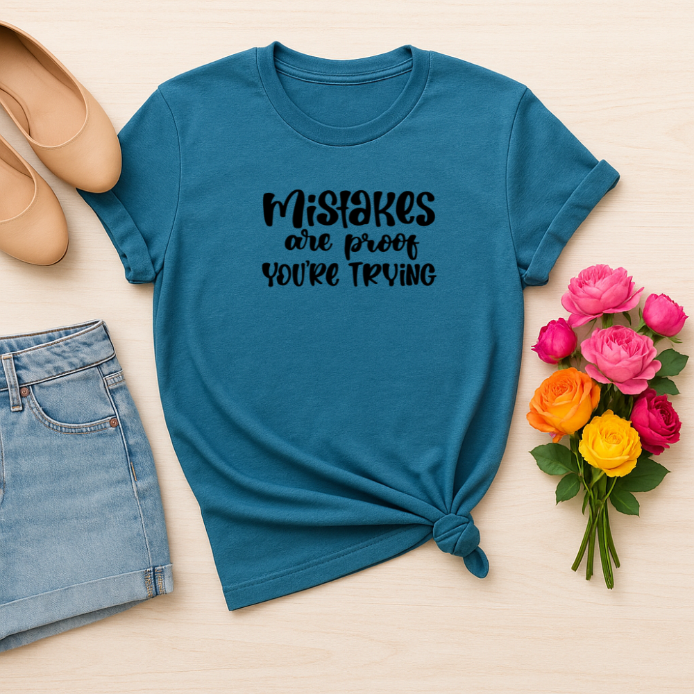 Blue t-shirt with 'Mistakes are proof you're trying' text, paired with jeans, shoes, and flowers on a beige background.