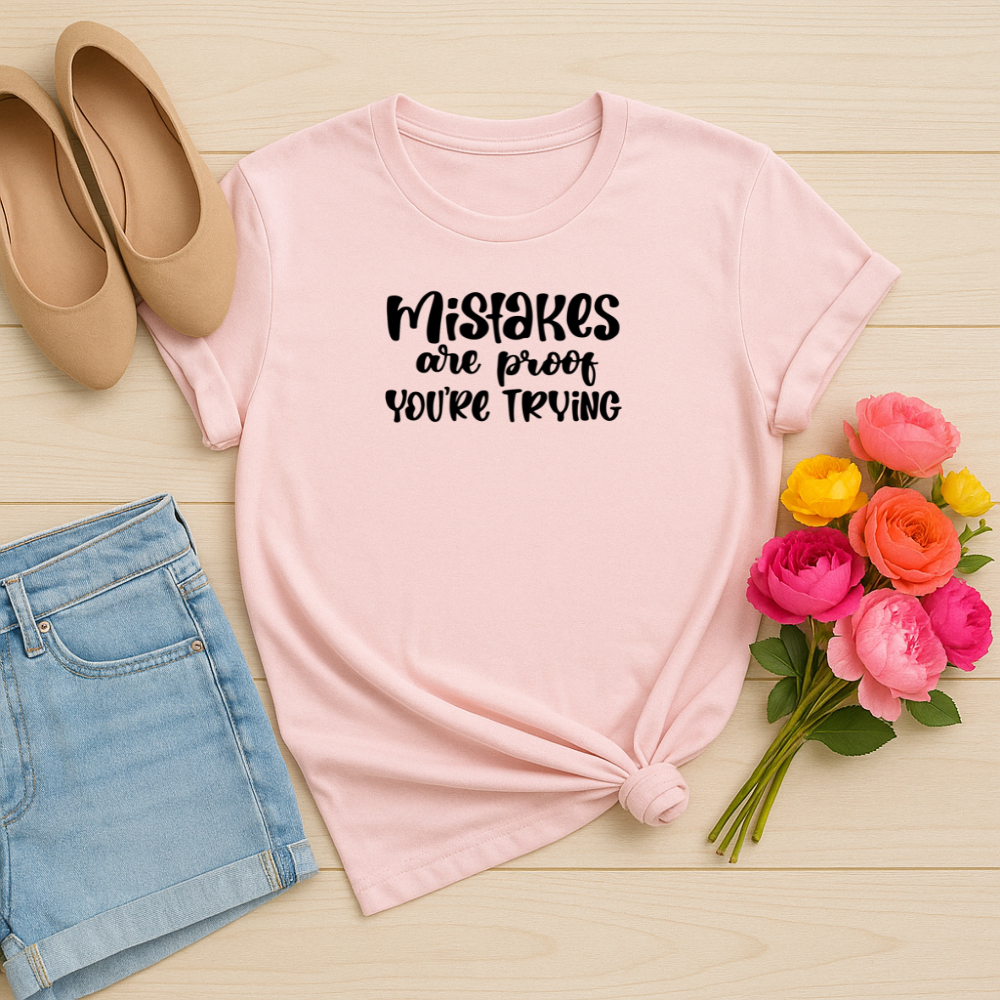 Pink t-shirt with 'Mistakes are proof you're trying' text, beige shoes, denim shorts, and flowers on a wooden surface