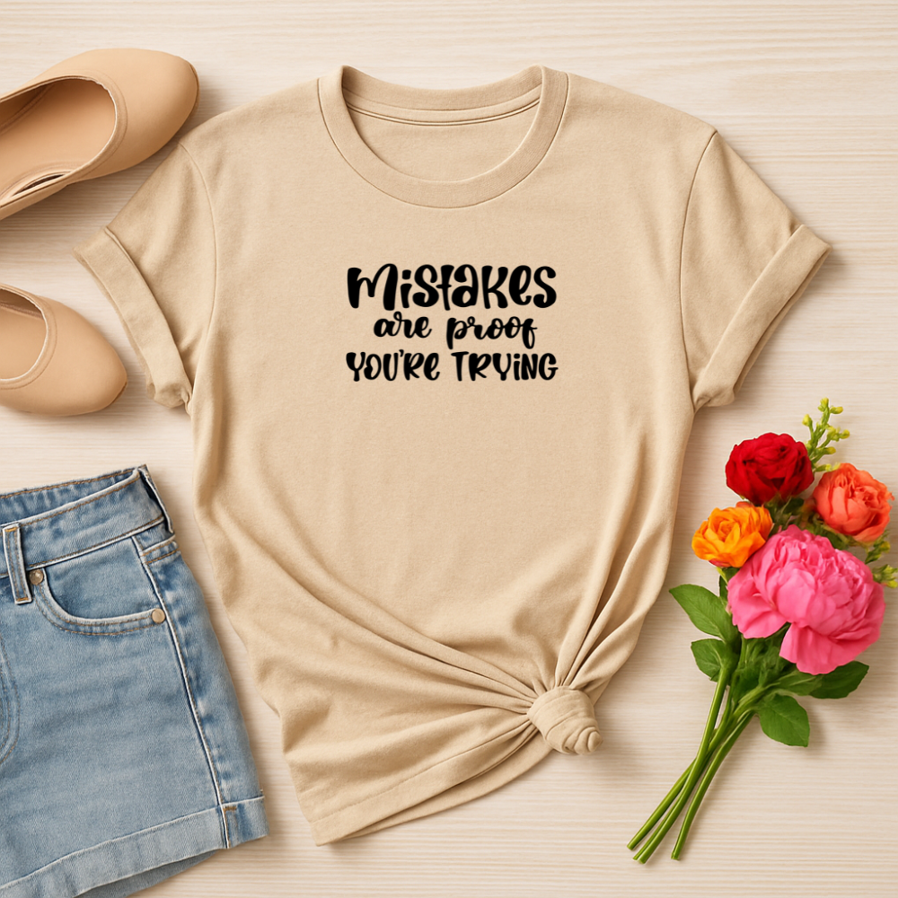 Beige t-shirt with 'Mistakes are proof you're trying' text, paired with jeans and flowers on a light wooden surface.