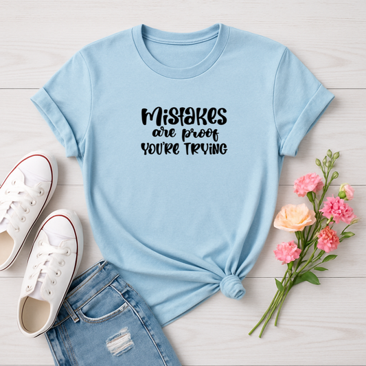 Light blue t-shirt with 'Mistakes are proof you're trying' text, paired with white sneakers and pink flowers on a light wooden surface.
