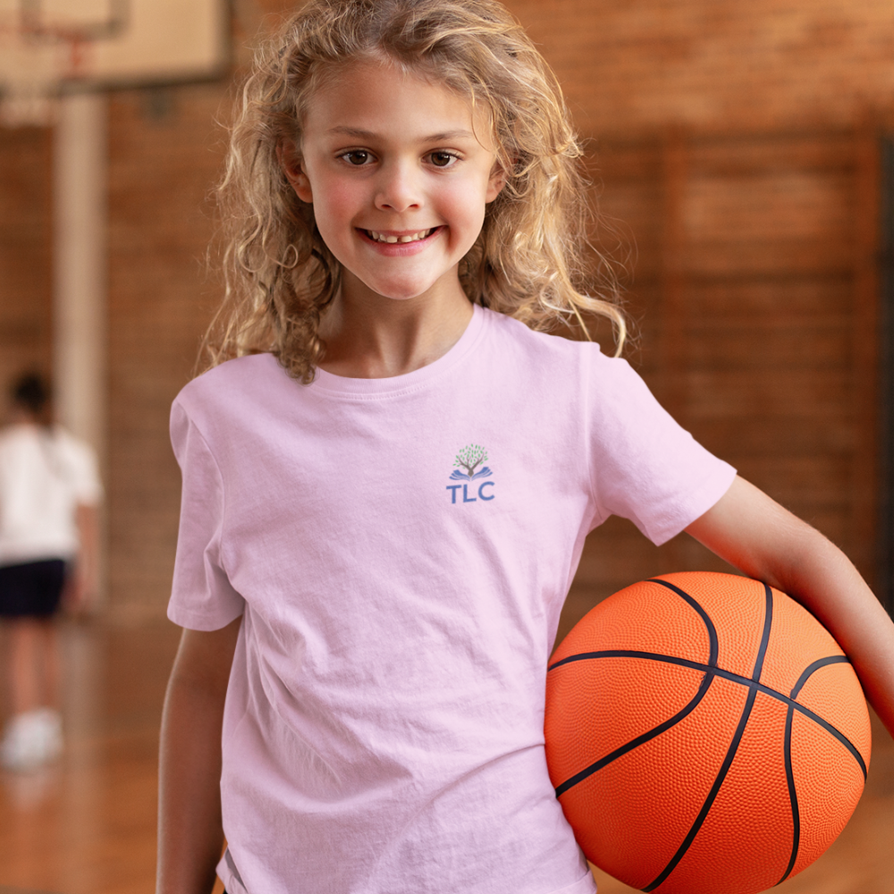 Young girl holding a basketball indoors, wearing a pink t-shirt with a logo.