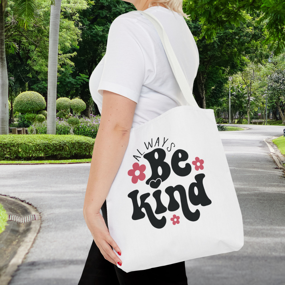Person holding a white tote bag with 'Always Be Kind' text in a park setting