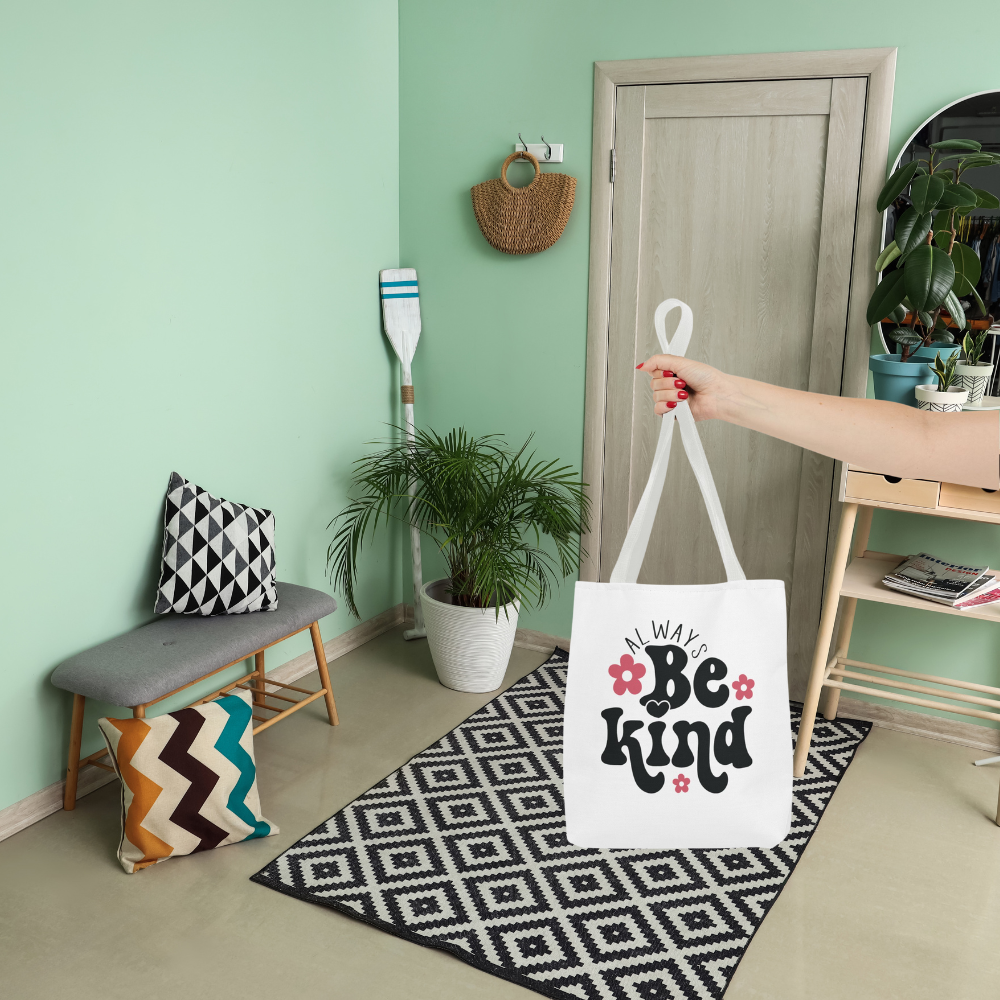 Tote bag with 'Be Kind' text held by a hand in a room with a green wall, bench, and plant.