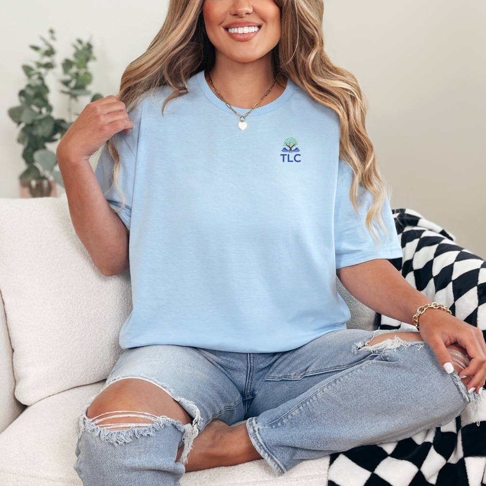 Woman wearing a light blue t-shirt with 'TLC' logo, sitting on a couch.