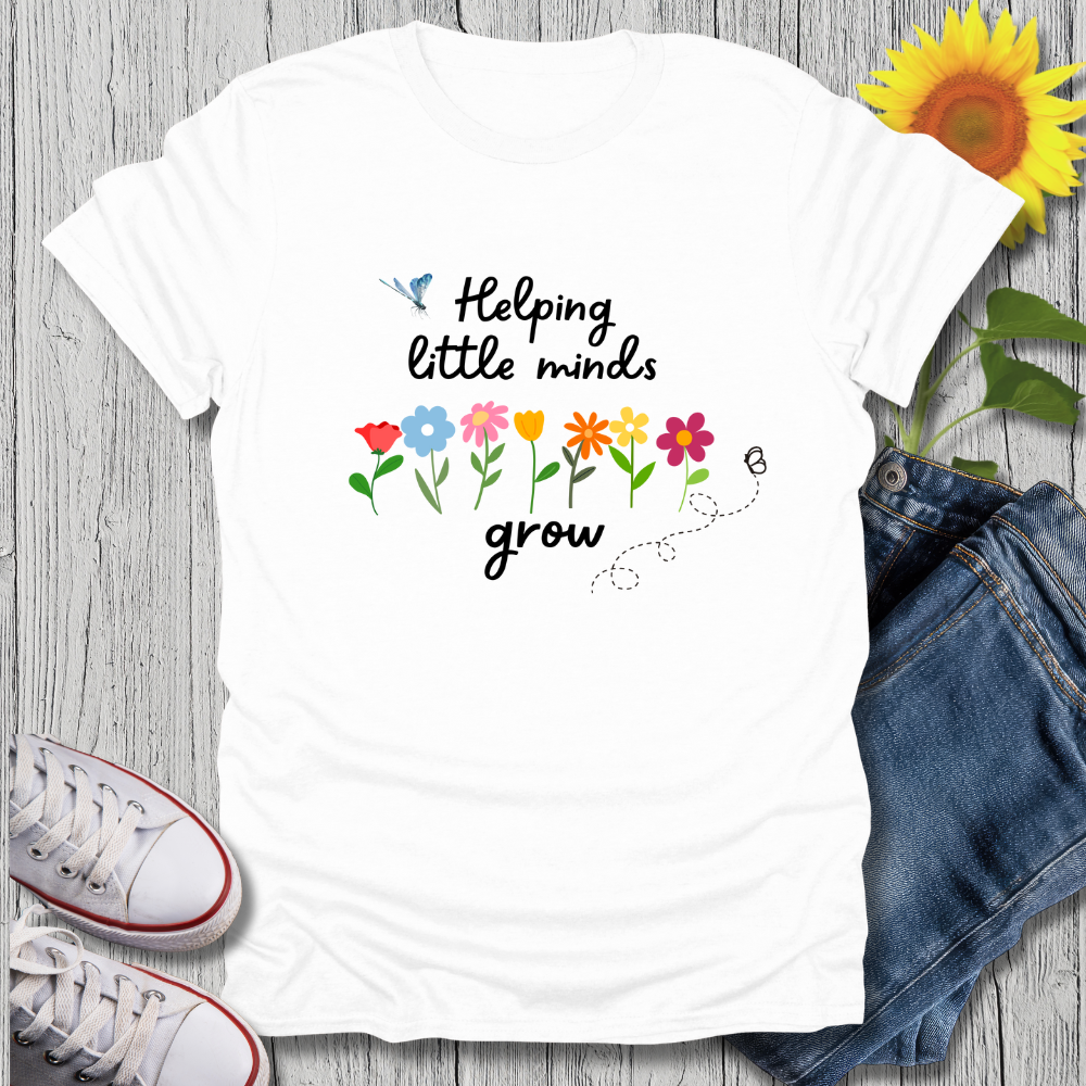White t-shirt with 'Helping little minds grow' text and flowers on a wooden surface with jeans and sneakers.