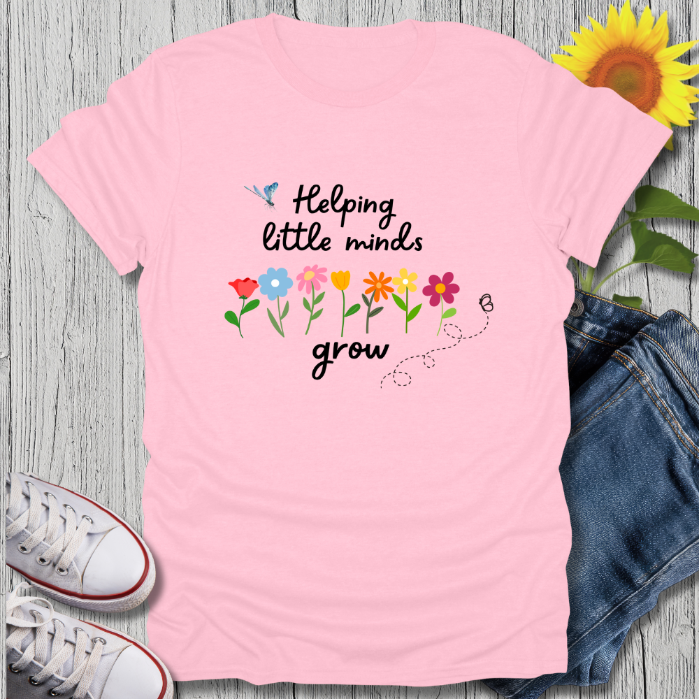 Pink t-shirt with 'Helping little minds grow' text and flowers on a wooden surface with jeans and sneakers.