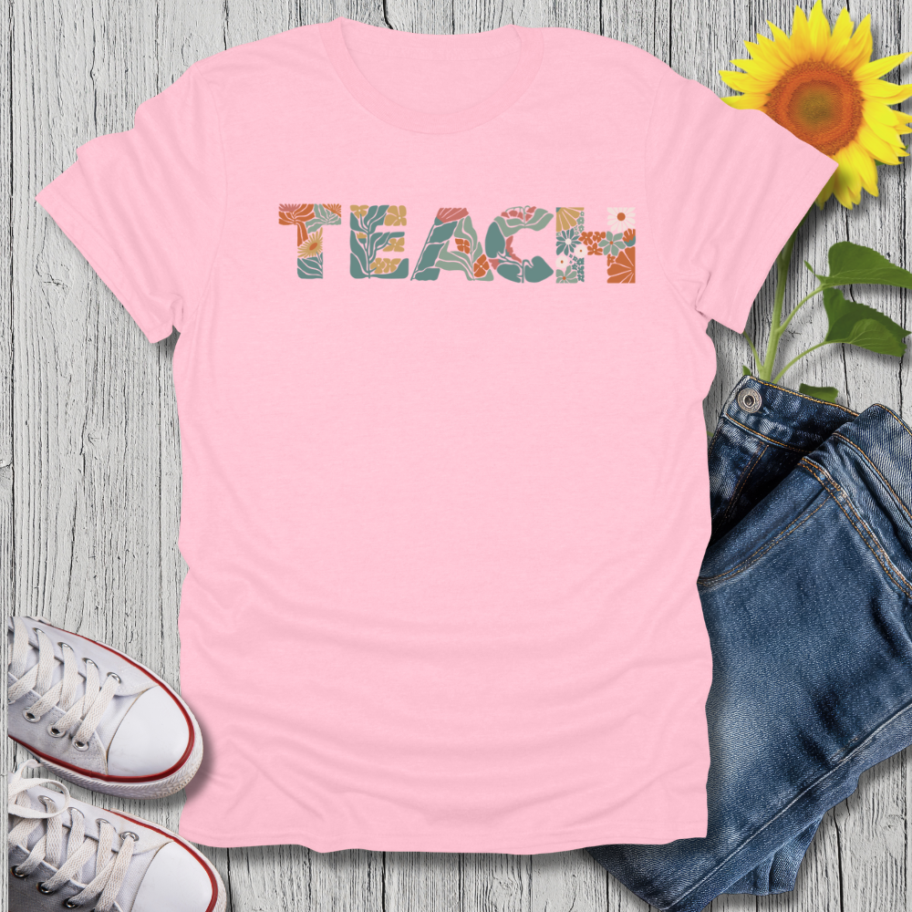 Pink t-shirt with 'TEACH' printed on it, next to jeans and sneakers on a wooden surface.