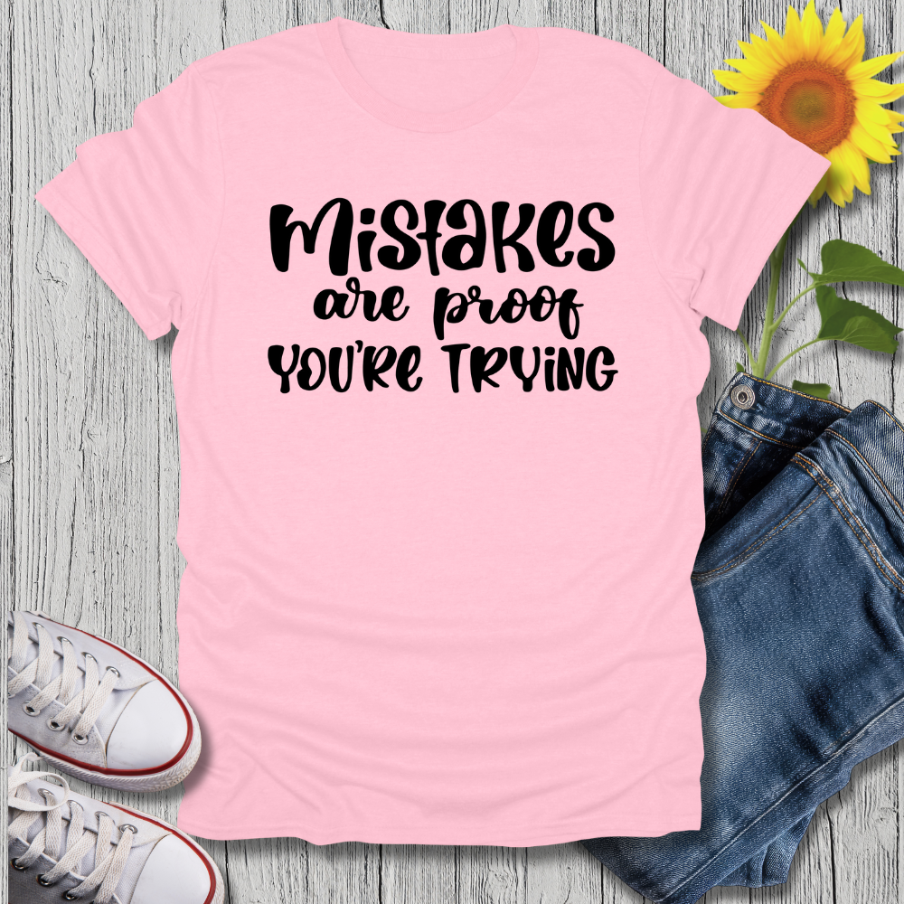Pink t-shirt with 'Mistakes are proof you're trying' text, paired with jeans and sneakers on a wooden surface.