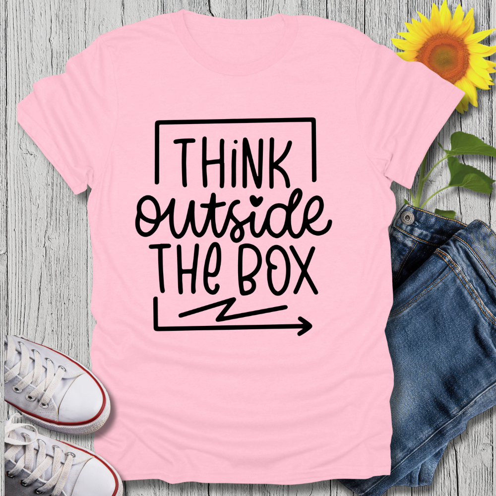 Pink t-shirt with 'Think Outside The Box' text, white sneakers, blue jeans, and sunflower on a wooden surface.
