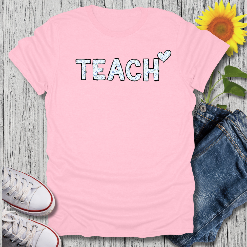 Pink t-shirt with 'TEACH' text, white sneakers, blue jeans, and a sunflower on a wooden surface.