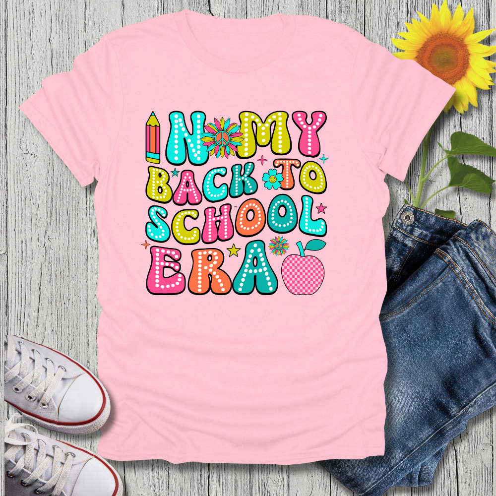 Pink t-shirt with colorful 'In My Back to School Era' text, jeans, sneakers, and a sunflower on a wooden surface.