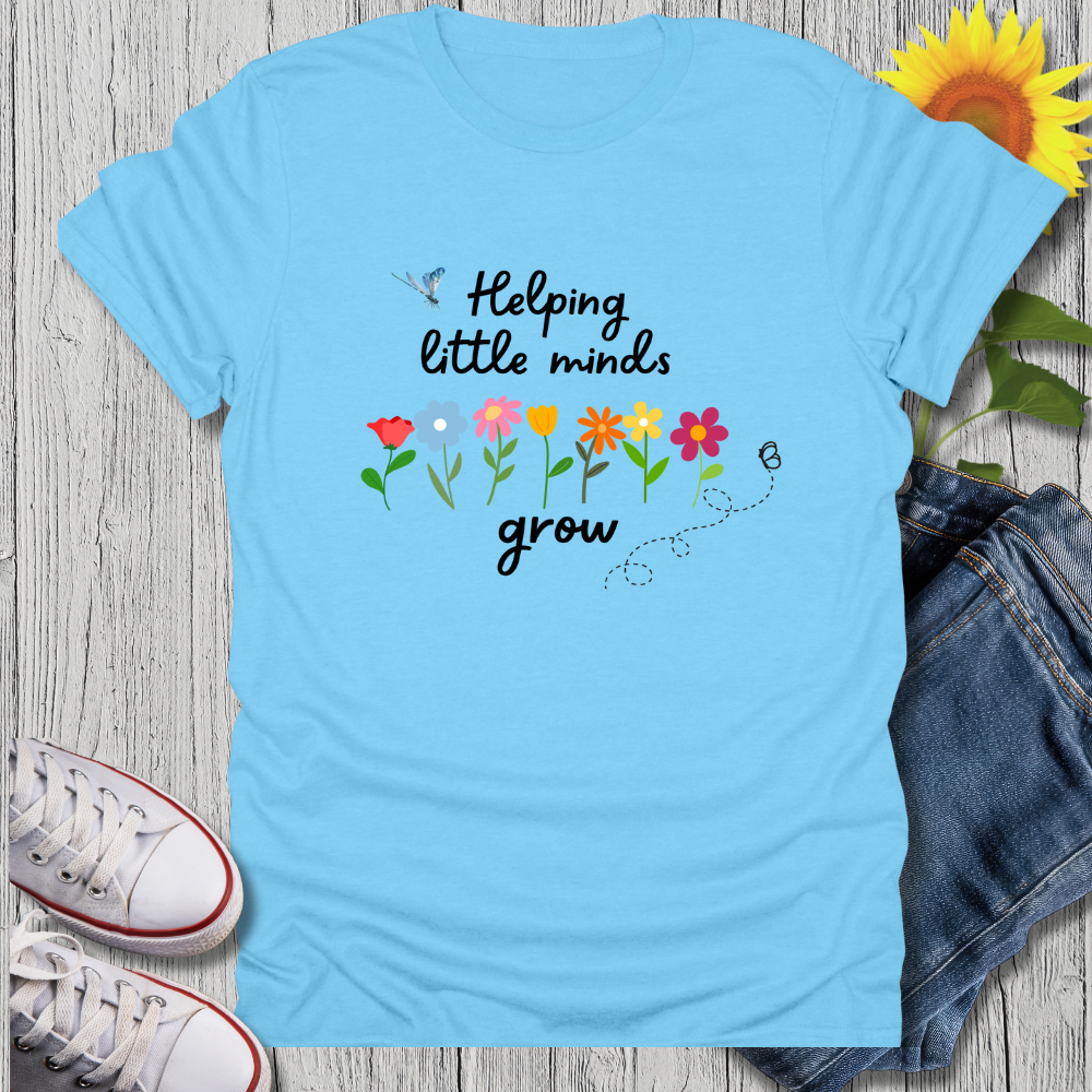 Light blue t-shirt with 'Helping little minds grow' text and flowers on a wooden surface with sneakers and denim shorts.