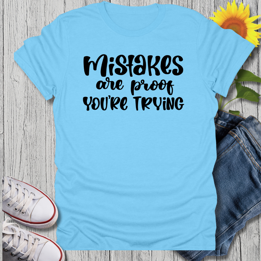 Light blue t-shirt with 'Mistakes are proof you're trying' text, paired with jeans and sneakers on a wooden surface.