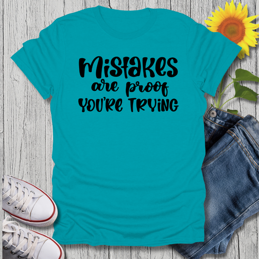 Turquoise t-shirt with 'Mistakes are proof you're trying' text, paired with jeans and sneakers on a wooden surface.