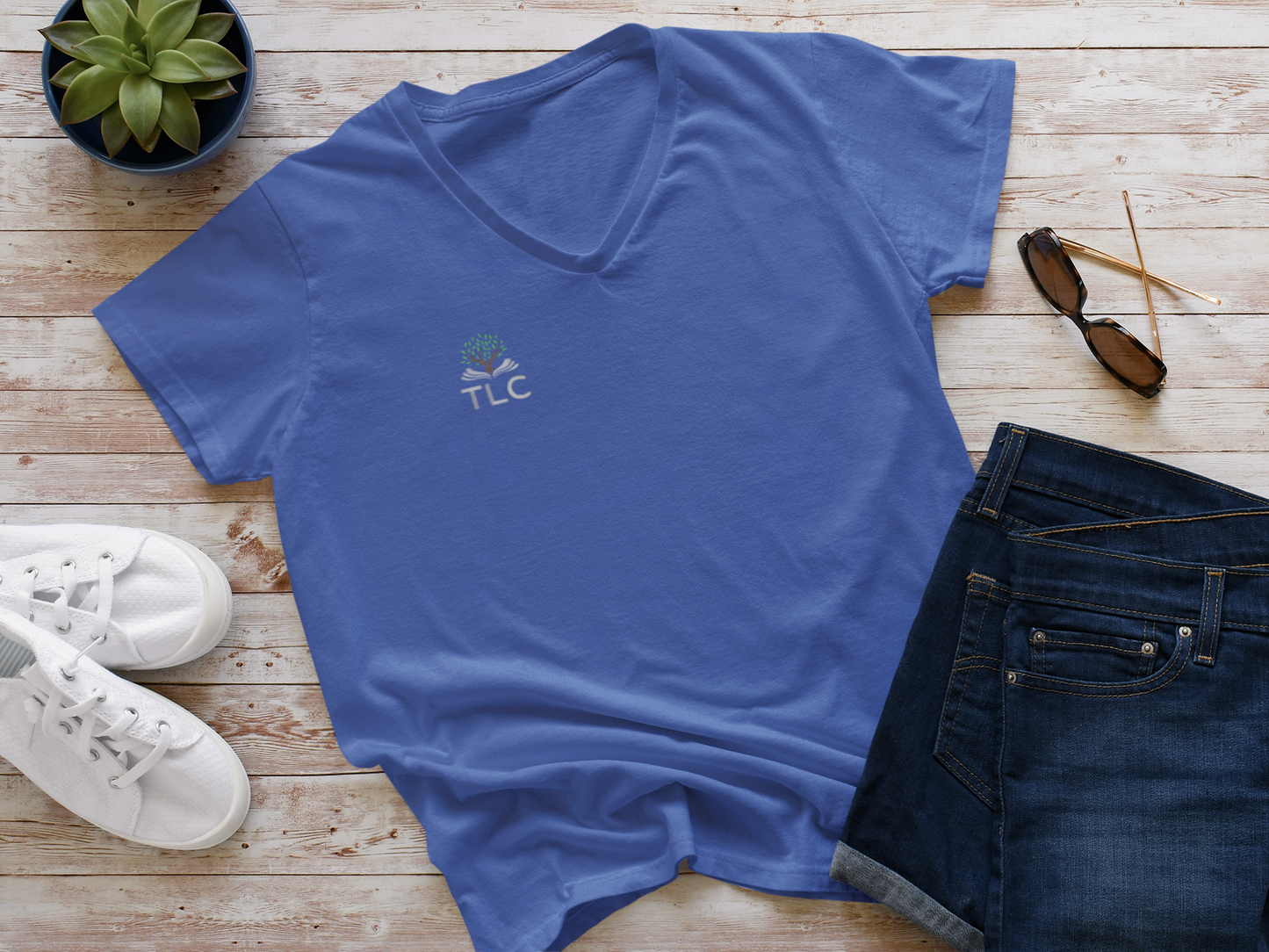 Blue t-shirt with 'TLC' logo on a wooden surface with jeans, sneakers, and sunglasses.
