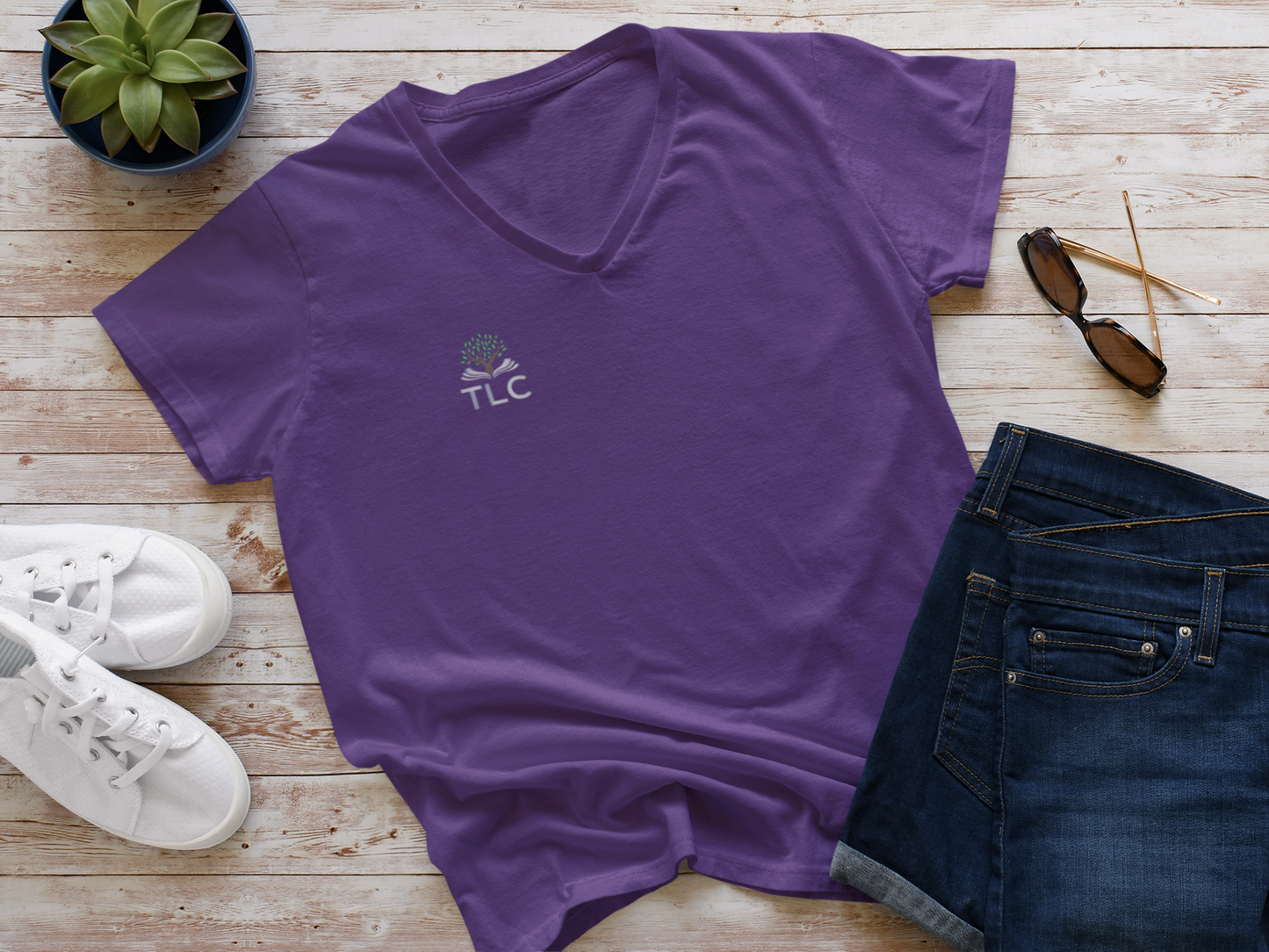 Purple t-shirt with 'TLC' logo on a wooden surface with jeans, sneakers, and sunglasses.